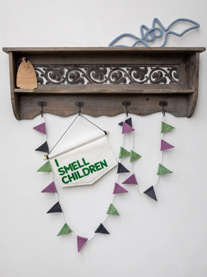 White, felt halloween banner that has "I SMEEL CHILDREN" written on it in green glitter felt. The banner is hanging from a wooden shelving unit surrounded by halloween decor.