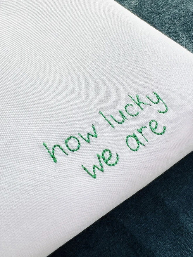 White embroidered T-shirt with the words “how lucky we are” in green block lettering