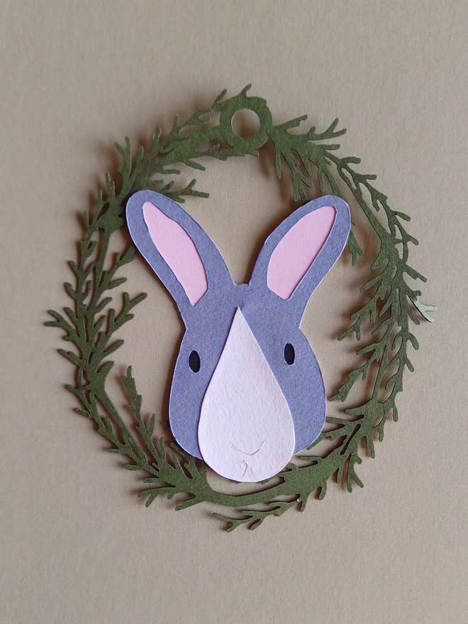 Black Bunny Easter Tree Decoration. Dark grey bunny face with white nose patch, black eyes and pale pink ears inside olive green foliage wreath oval