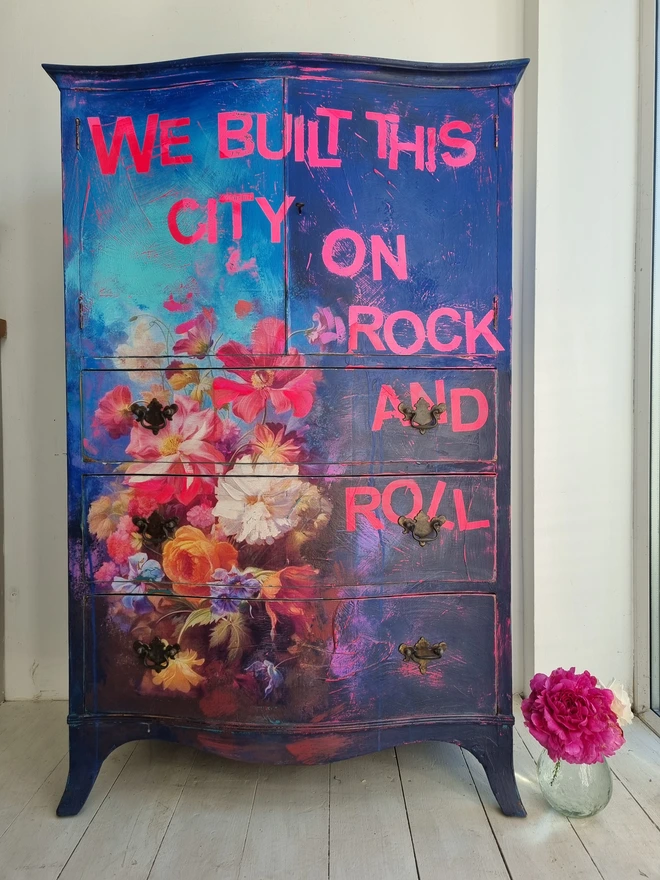 A vintage tallboy cabinet painted in vibrant blues, neons and flowers with the text that reads 'we built this city on rock and roll' in neon pink.