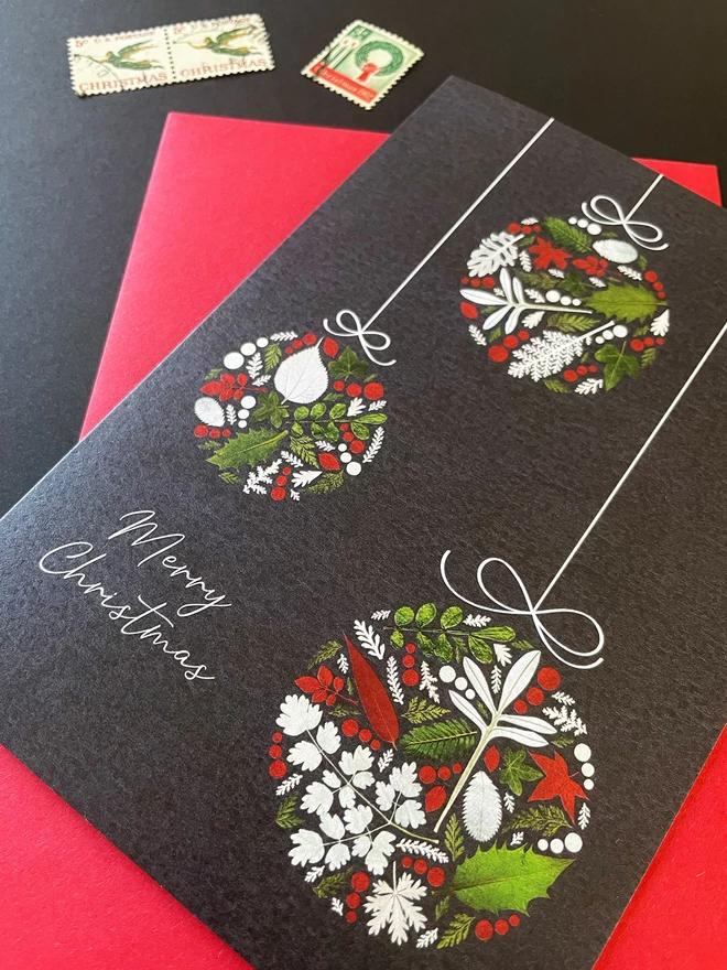 Close-Up of Christmas Card with Pressed Winter Leaves Bauble Design, Holly, Ivy, and Red Envelope on Dark Charcoal Desk