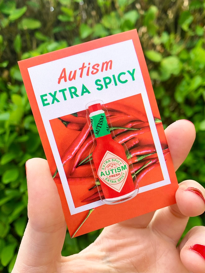 image shows a hand holding a red edged business card with the words ‘Autism Extra Spicy’ printed across the top. Pinned to the card is an acrylic badge in the shape and colours of a tiny hot sauce bottle. The bottle is labelled to say ‘fresh autism’. 