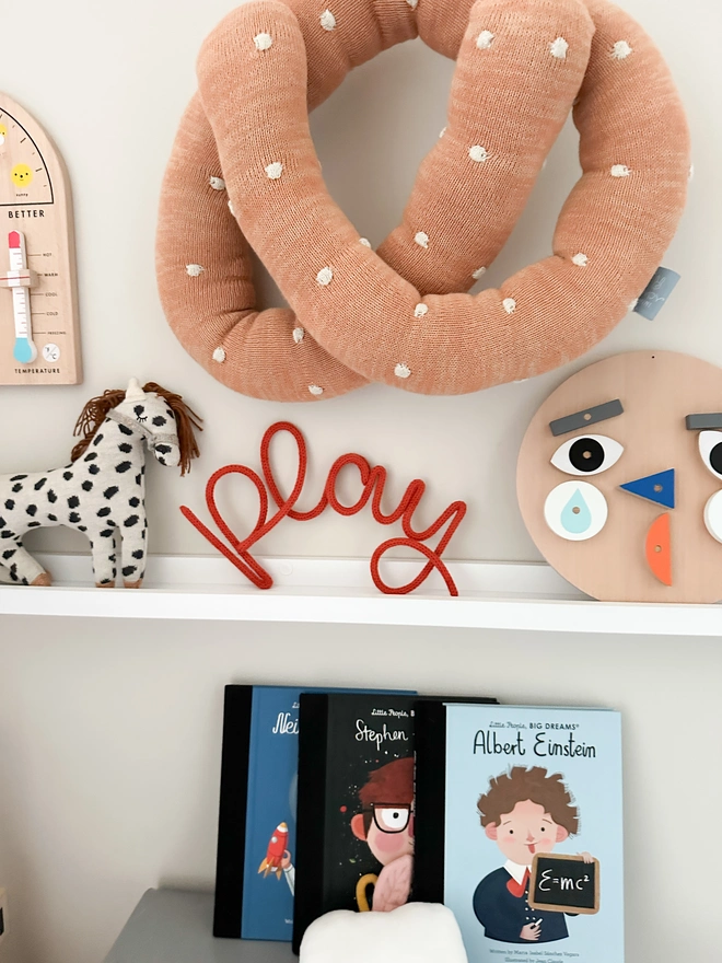 A 'Play' knitted wall sign on a shelf in a kids playroom or bedroom with other kids wooden toys, a soft toy horse and some childrens books all decorating the child's room. 