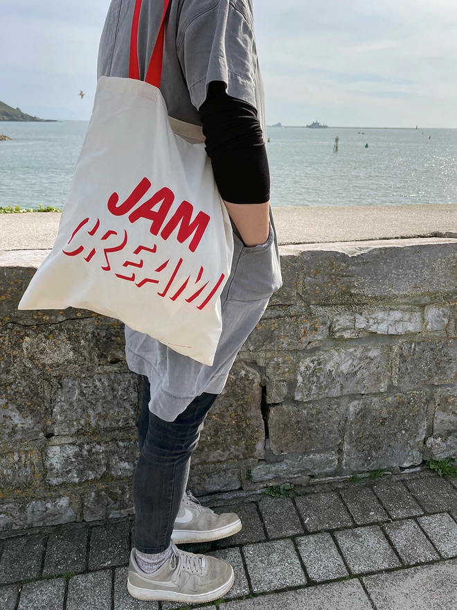 Woman in grey, stood looking out to sea, with a tote bag over her shoulder with JAM written above CREAM in red ink screenprinted on cream bag with red handles.