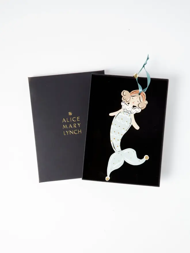 Mermaid Tree Dancer. the mermaid tree dancer hanging ornament in pale blue and gold tones sits inside its black presentation box, with the lid open to reveal "alice mary lynch" embossed in gold lettering. the mermaid figure has curled hair and an orange headband, with a pale blue scalloped scale-patterned tail curling into a jester-style pointed tip with gold-tipped ends. the branded box gives the piece a polished, gift-ready presentation.