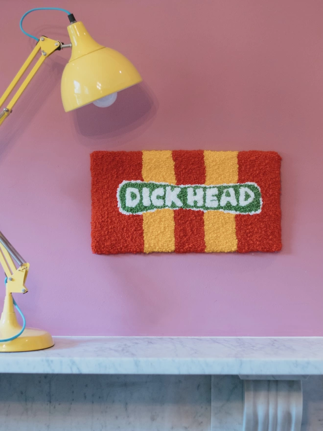 'Dick Head' Handmade Tufted Rug/Wall Hanging seen hanging on a pink wall above a mantlepiece with a yellow lamp.