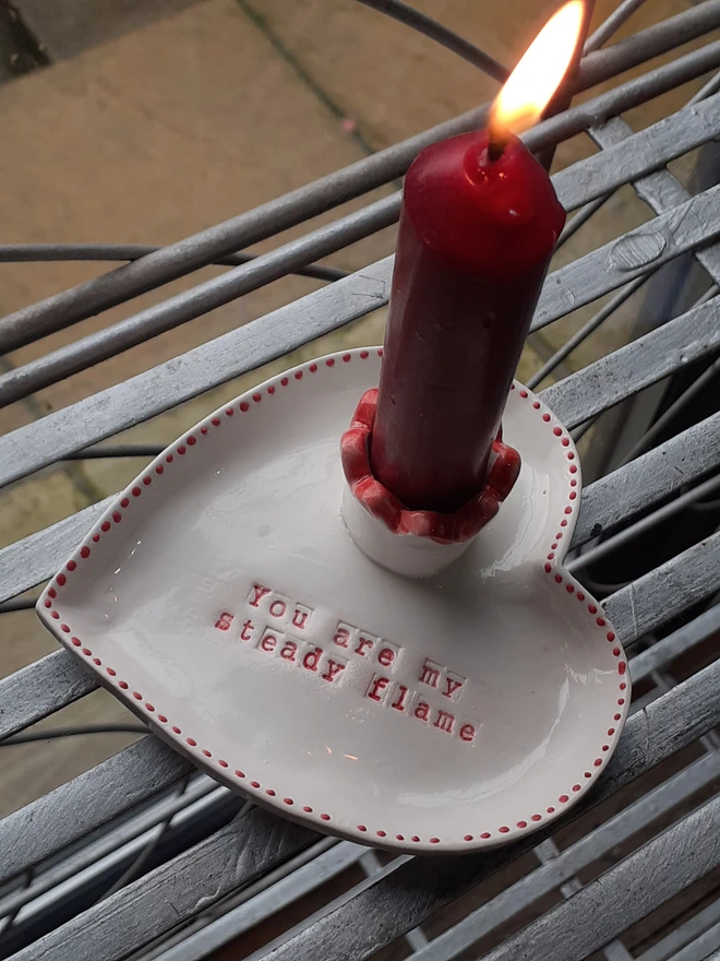 Handmade ceramic Valentine candle holder shaped like a white heart with red dotted border, holding a lit red taper candle and stamped with the words ‘You are my steady flame’