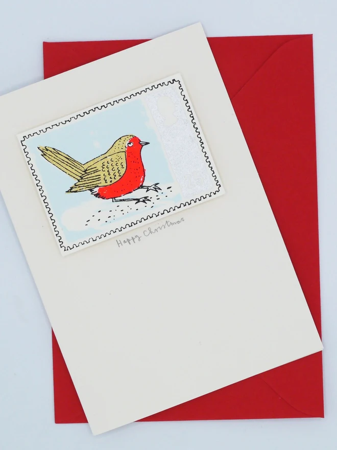 Robin Stamp Christmas Card, a white Christmas card with a colourful bird design in the centre on top of a red envelope on a plain surface. 