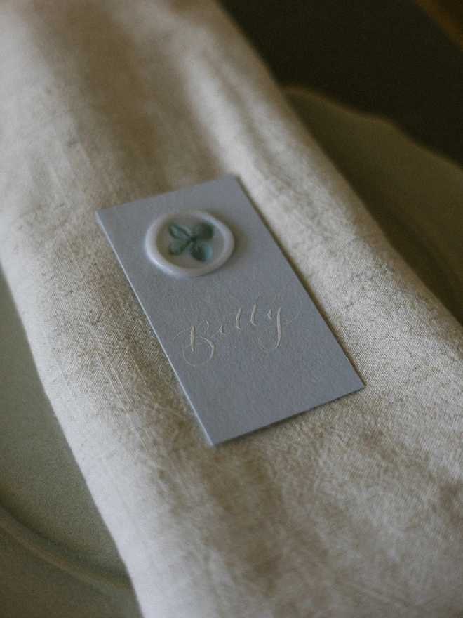 Pale blue place card with white calligraphy name and white vellum wax seal with hydrangea petals on a linen napkin 