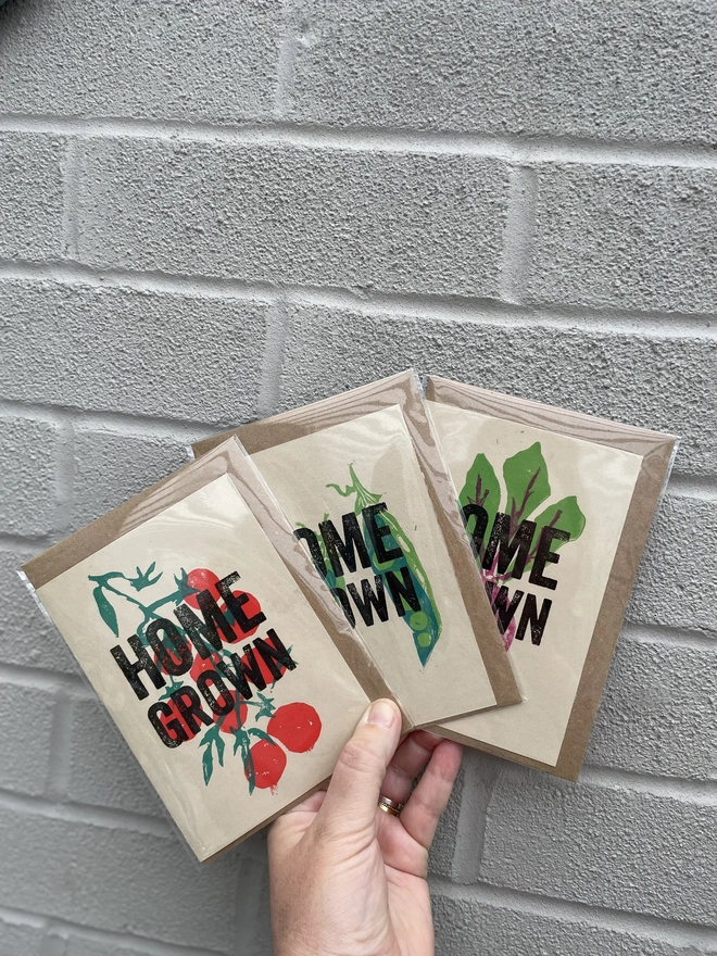 'Home Grown' Tomatoes Lino Garden Print.  A display of handmade tomato-themed cards arranged in a hand against a brick wall. 