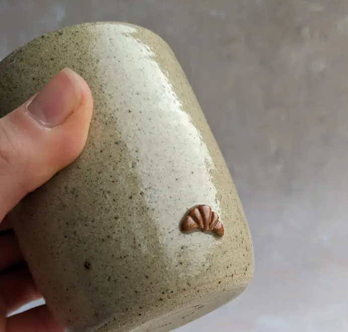 Biscuit coloured croissant coffee cup with a subtle croissant design, placed against a grey marble background.