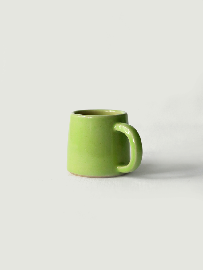 A handmade ceramic espresso mug in spring green glaze, with a chunky loop handle, photographed against a plain light grey background
