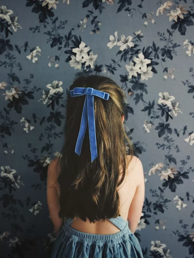 Child standing in front of blue floral wallpaper wearing the blue velvet hair bow.