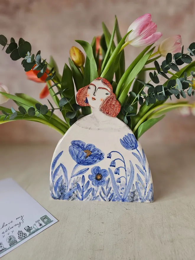 a large vase shaped like a lady with red hair looking up with blue flowers painted on her dress, front view with flowers