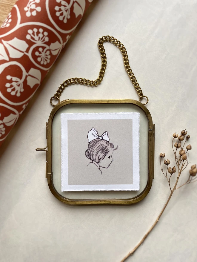 a photograph of a miniature hand drawn portrait of a little girl with a big white hair bow. framed in a small glass frame with a brass edge