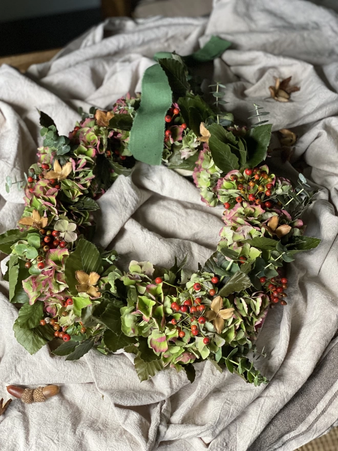 A portrait shot of a full handmade Fresh Rose Hip And Hydrangea Wreath with berries and eucalyptus, on display with a sage green ribbon looped around the wreath, atop a soft grey ruffled fabric