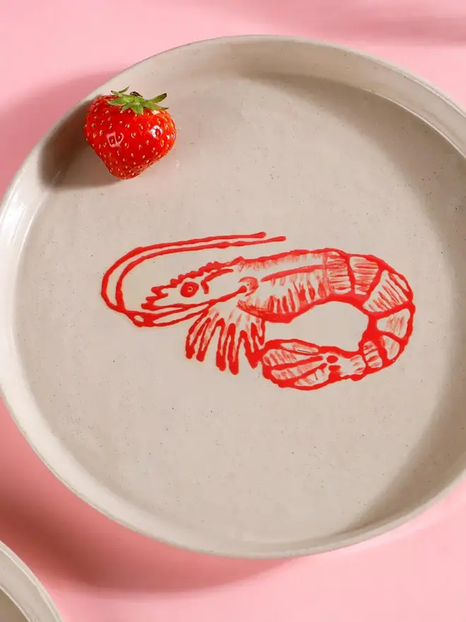 Handmade Ceramic Side Plate With A Red Prawn Design, a plate with a red prawn design sitting on a pink table surface. 