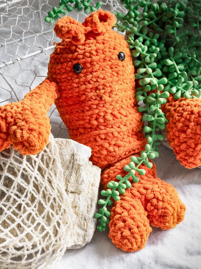 Chunky Chenille Cuddly Crochet Crustacean Lobster Plush, an orange lobster plush sitting accompanied by a bag and some greenery. 