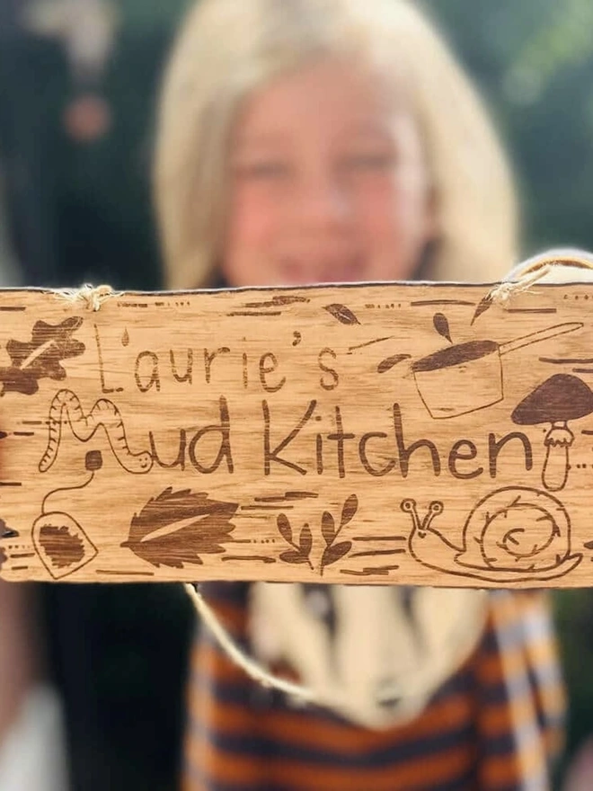 Personalised Wooden Mud Kitchen Sign, a child holding a personalised wooden mud kitchen sign outdoors. 