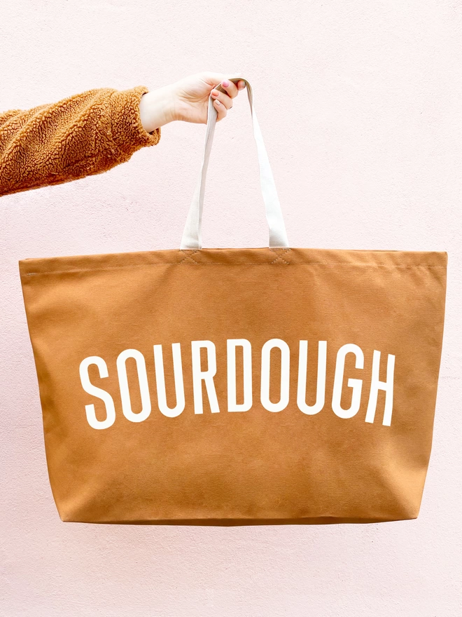 model holding out an oversized tan canvas tote bag with sourdough slogan on a pink background