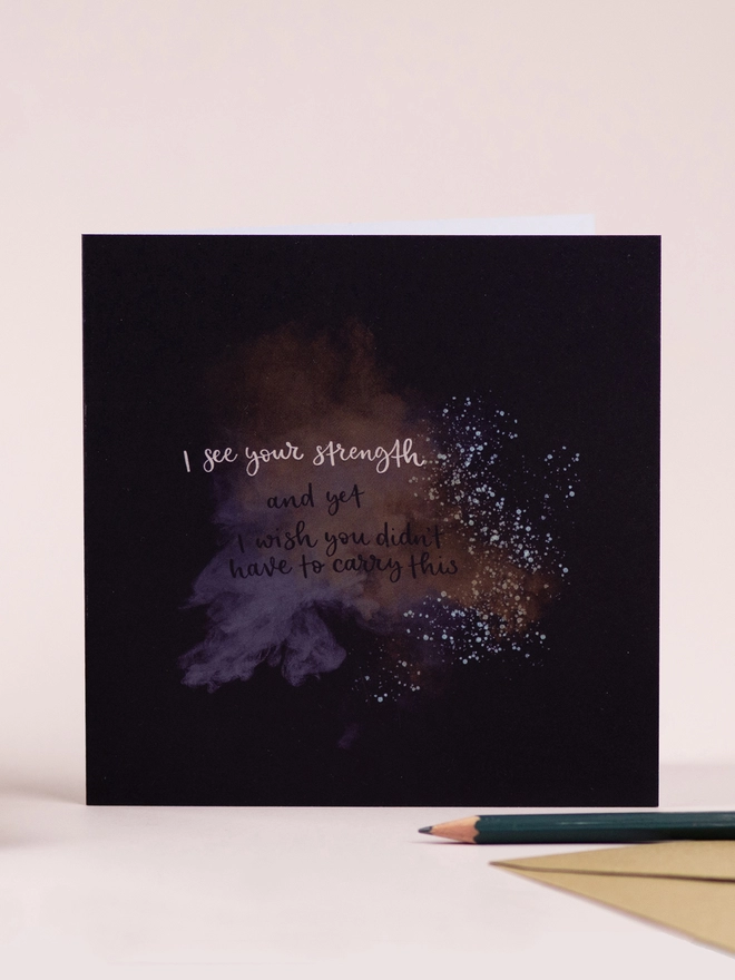 A deep purple card stands against a pale pink backdrop, with a pencil and a brown envelope just visible. The card features an abstract powder-burst design in ochre and purple.