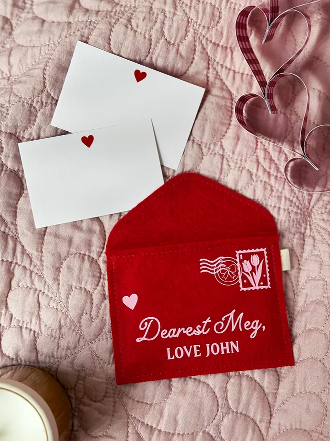 A handmade red felt envelope with a pale pink stamp and postmark design, and the words 'Dearest Meg, love John' rests on a pink quilt. Two notecards with small hearts on are tucked under the top of the envelope.