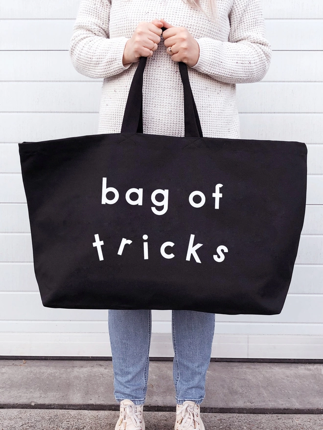 model holding out an oversized black canvas tote bag with the words bag of tricks