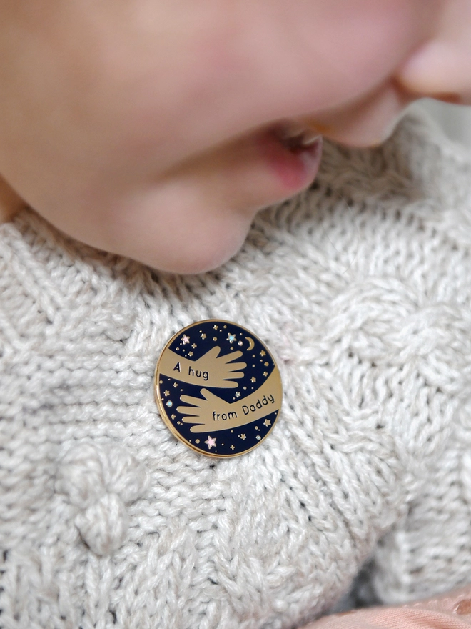 A navy blue and gold enamel pin badge with a hugging arms design and the words "A hug from Daddy" is pinned to a young child's cardigan.
