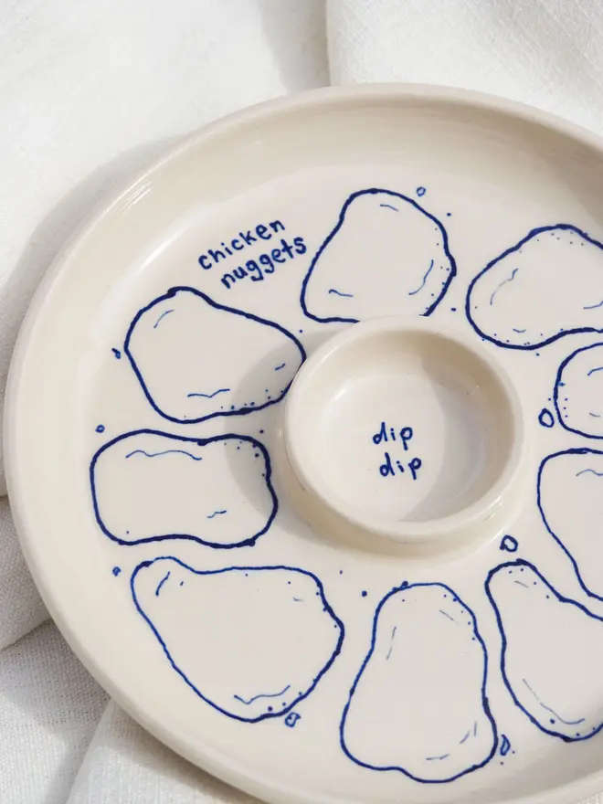 Snack Chicken Nuggets & Dip Plate, a white snack dish sitting on a white bed cloth. It is covered in blue outlines for chicken nuggets. 