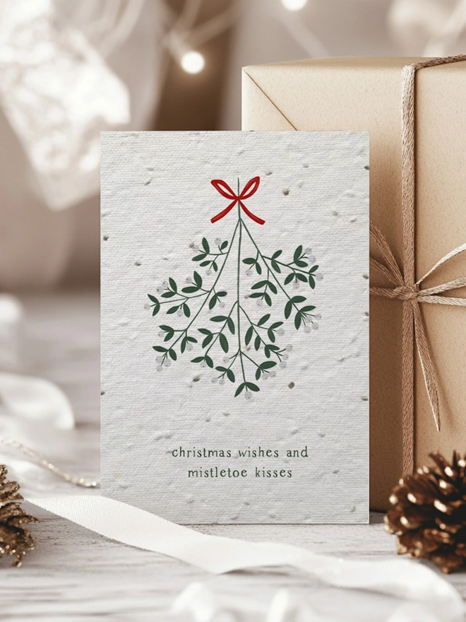 Christmas Card with illustration of mistletoe with 'Christmas Wishes and Mistletoe Kisses'. In the background is Christmas presents, greenery and pine cones.