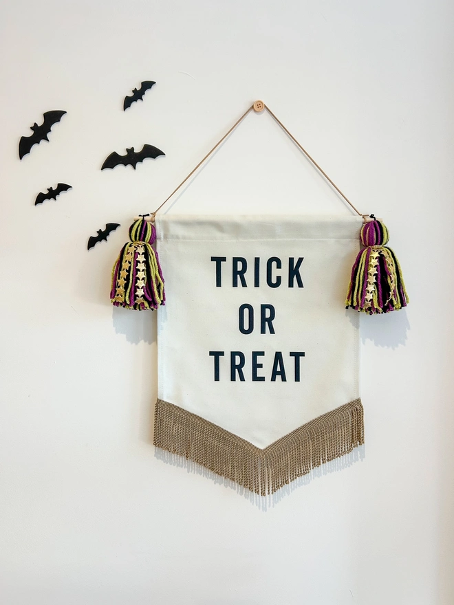 Trick Or Treat Halloween Fabric Banner with purple, black and green tassels with gold star detail and gold bullion trim. Hanging on a white wall with black bats 