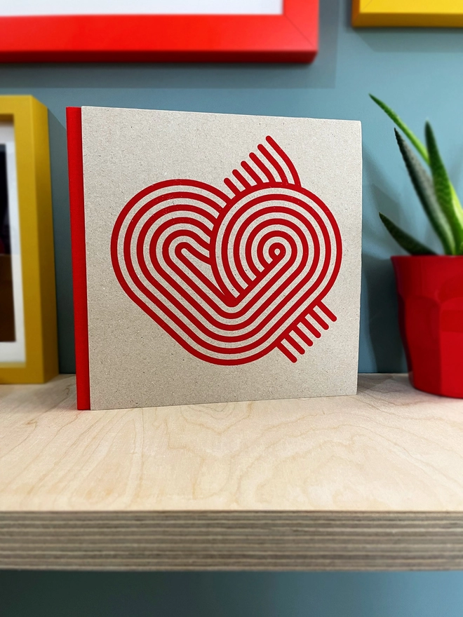 Stripy Heart design is screenprinted in magenta on a grey pasteboard sketchbook with a red fabric spine, stood on a plywood shelf with hints of framed pictures around. the wall is duck egg blue, a plant glimpsed to the side.