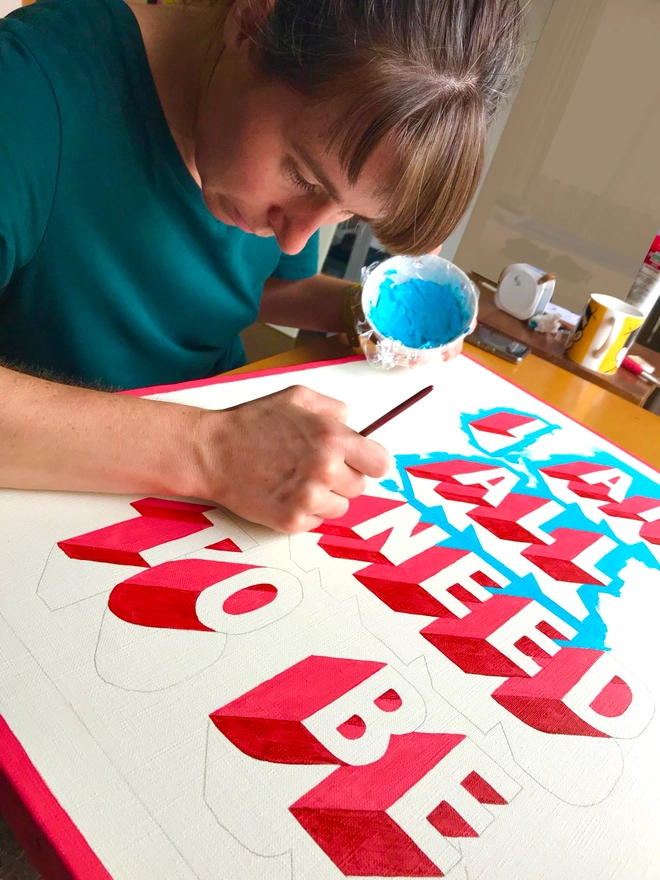 Artist Survival Techniques painting a partially finished typographic artwork that reads “I AM ALL I NEED TO BE.” The artist is leaning forward, carefully applying blue paint to one of the bold 3D letters. The text features red and white lettering with blue shadows on a blue background.