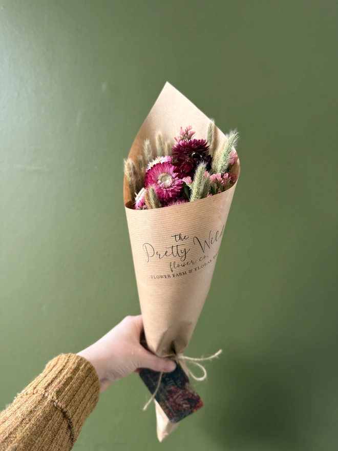 a bunch of dried flowers in bright colours wrapped in kraft paper held by a person in a mustard coloured jumper with their left arm against a green wall