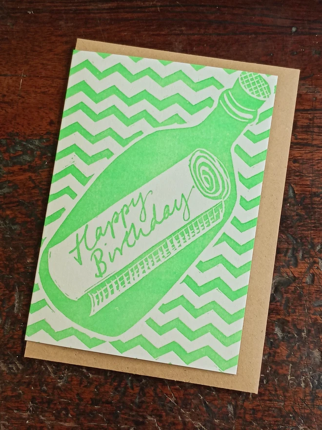 Green card with envelope folded inside. Printed on front: Flat green, cartoon style. Green zigzag pattern backing matte bottle outline containing scroll with printed text, "Happy Birthday"