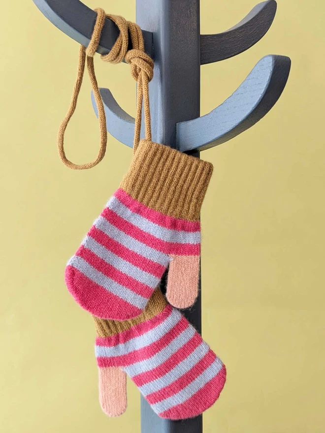 Kids Pink And Light Blue Stripe Mittens On A String Hanging On A Coat Stand In Front Of  A Green Background