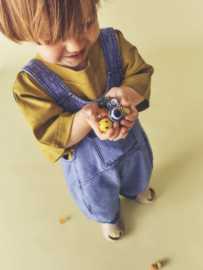Toddler smiles standing among scattered marbles against a pale yellow backdrop, wearing Studio Koter washed blue denim Kai dungarees over an ochre t-shirt.
