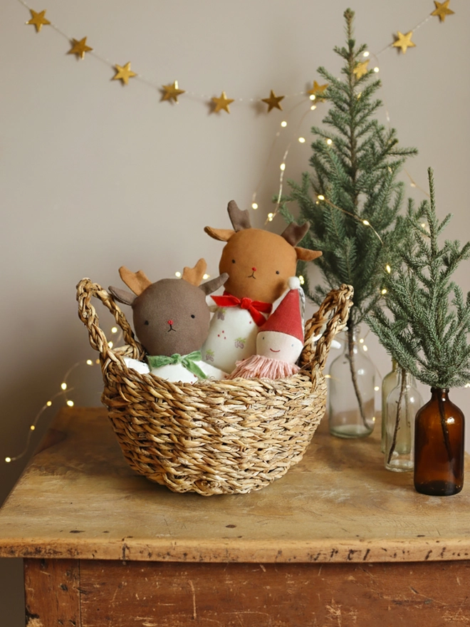 Two handmade reindeer dolls in a basket. Each doll has soft brown antlers, embroidered facial features, and wears a festive white outfit with a pinecone print. One doll has red legs and a red velvet bow; the other has green legs, a green bow. 