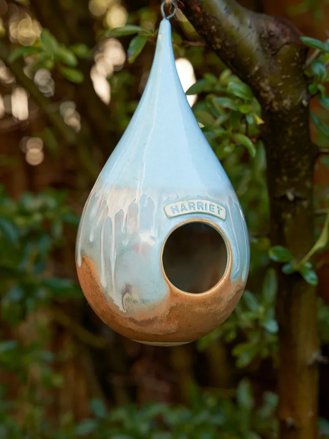 Personalised blue teardrop bird feeder hanging from a tree personalised with the name 'Harriet'