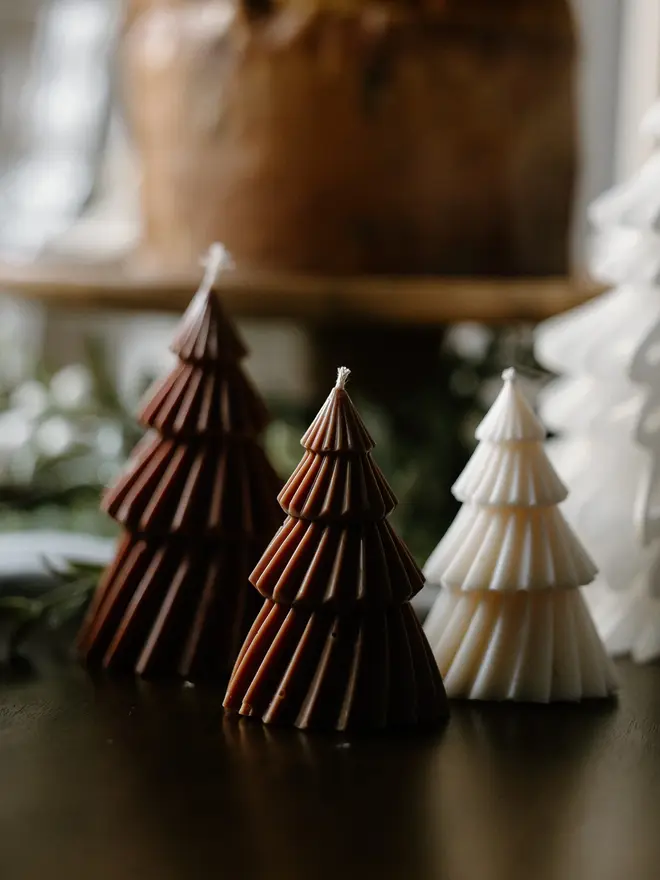Brown Christmas Tree Warm Candles (Set Of 2). Brown and cream tree-shaped candles with crisp pleated tiers lined up on a dark table, with greenery and a wooden board blurred behind. The mix of colours gives a cosy, Scandi-inspired look to festive décor.