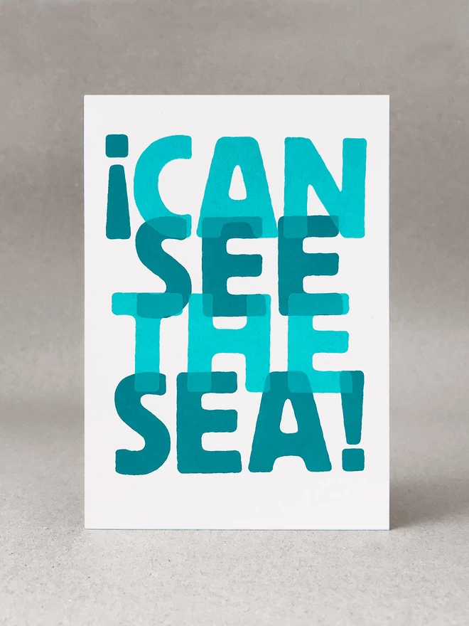 I can see the sea, screenprinted words overlapped in two turquoise blues, on a white card. It is sat in a light grey studio set.