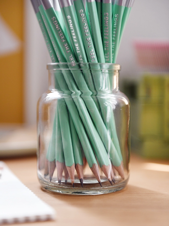 A glass jar full of green pencils with silver tips stands on a wooden desk. Several stationery items lay beside it.