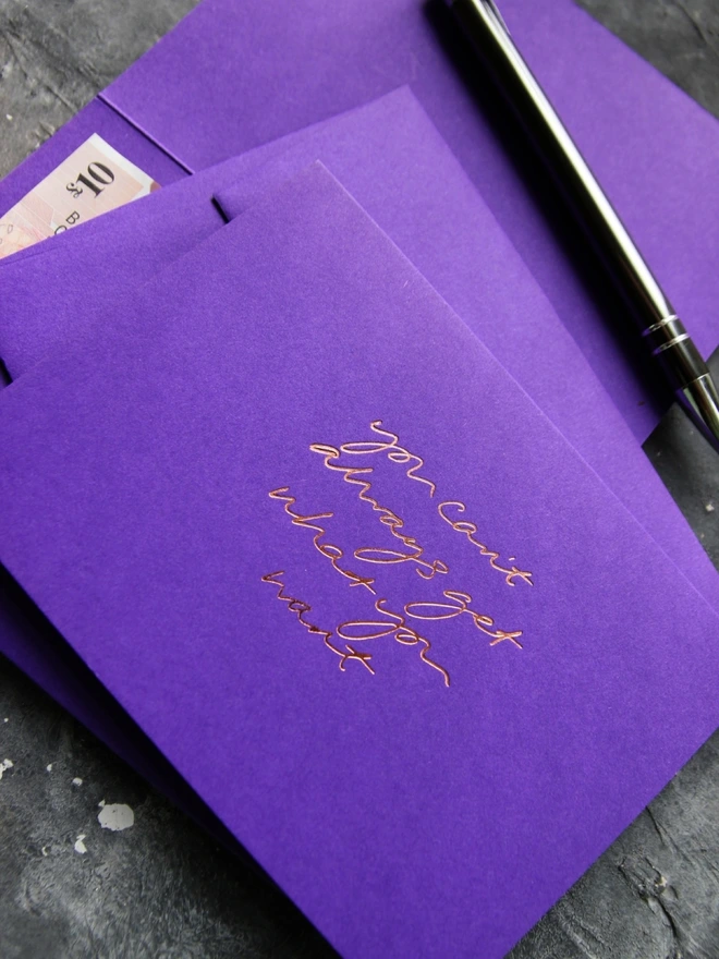 Hand foiled cash card or money wallet in a royal purple colour handfoiled in shiny rose gold foil text which says ‘You Can’t Always Get What You Want’.