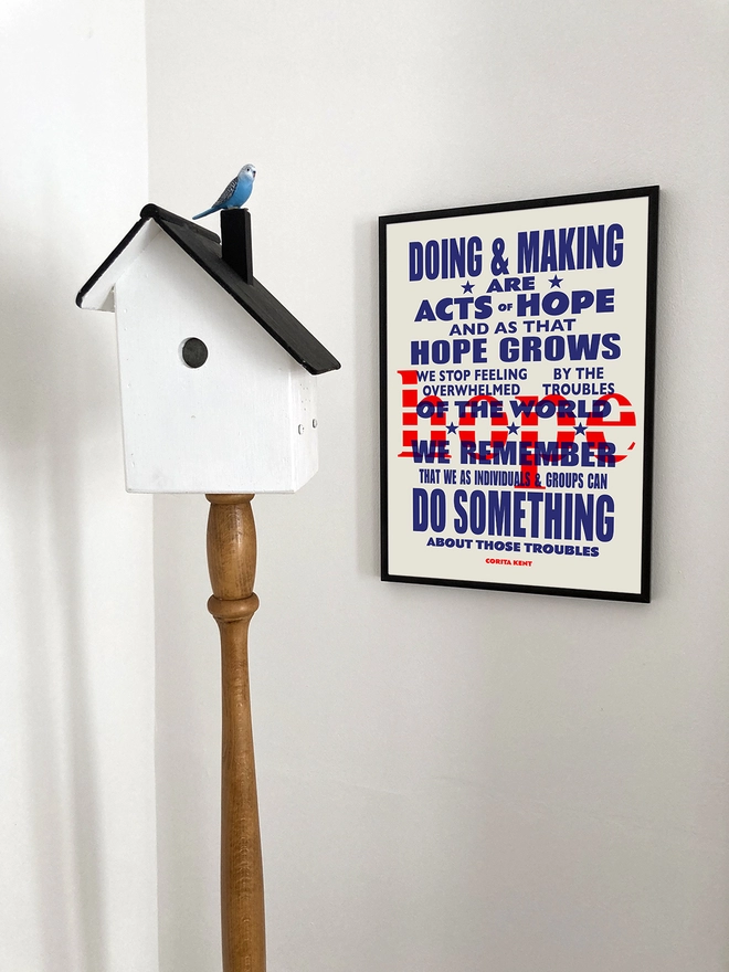 Framed multicoloured typographic print of “Acts Of Hope” The print hangs next to a bird box standard lamp with a small blue budgie sitting on the chimney.