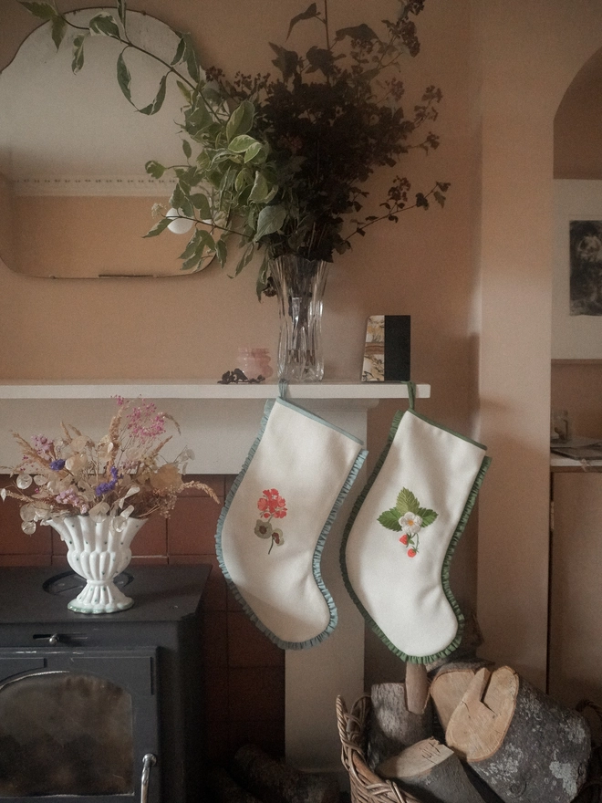 Two ivory embroidered Christmas stockings with geranium and strawberry botanical motifs, hanging beside a wood-burning stove and basket of logs in a cosy country home.