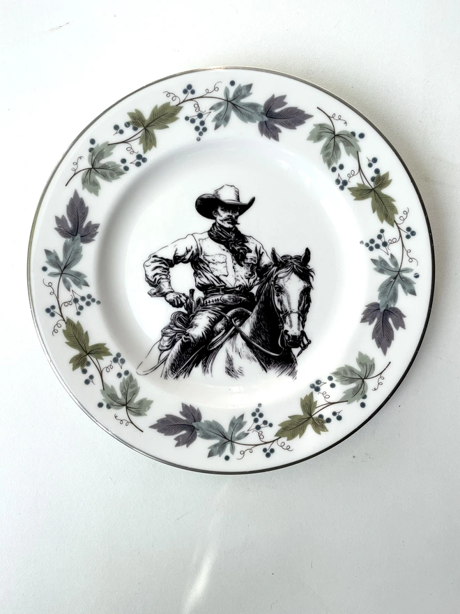 vintage plate hanging on a white wall, it has a decorative border of grey and blue leaves, with a printed vintage illustration of a cowboy in the middle 