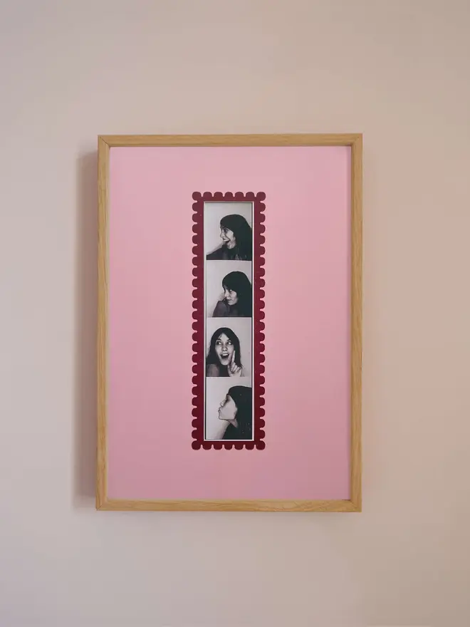 Oak A4 picture frame with a pink mount and scalloped red border, displaying a centered vertical photo booth strip, hung on a neutral beige wall.