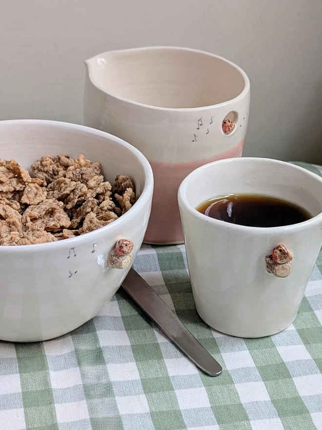 white ceramic bowl with a tiny modelled robin perching on a modelled branch with musical notes, there is a pink ceramic jug with a robin bird and a white espresso cup also with a robin all on a green gingham tablecloth