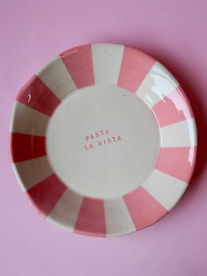Handmade ceramic pasta bowl with pink striped rim and the words “Pasta La Vista” stamped in the centre, photographed on a pink background.