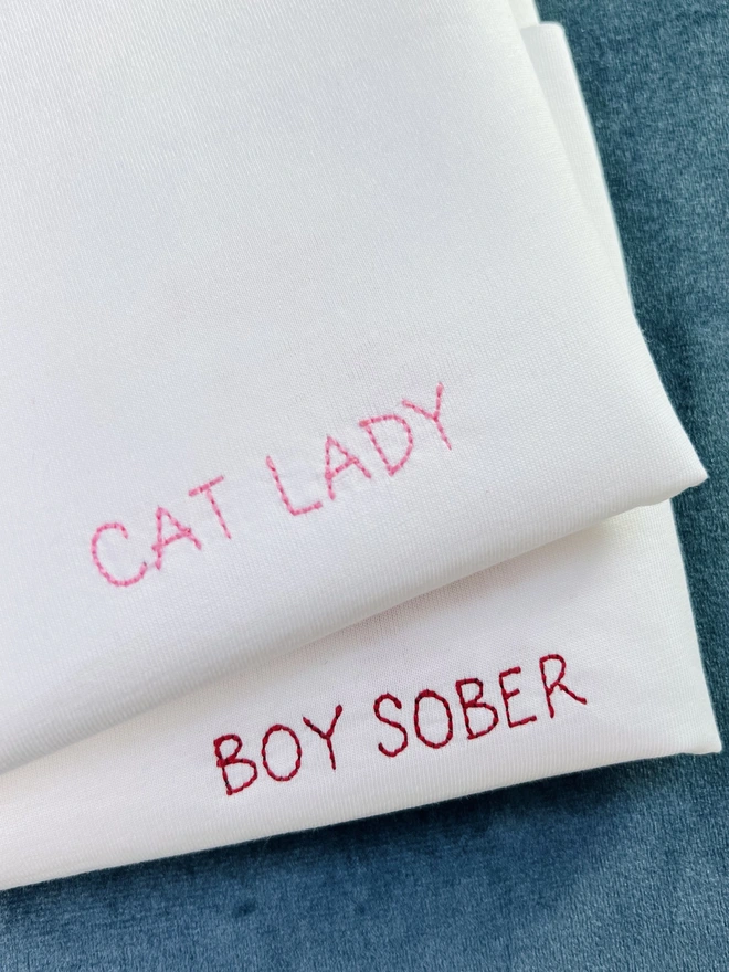 Two white embroidered T-shirts with pink and red personalised text reading “Cat Lady” and “Boy Sober”